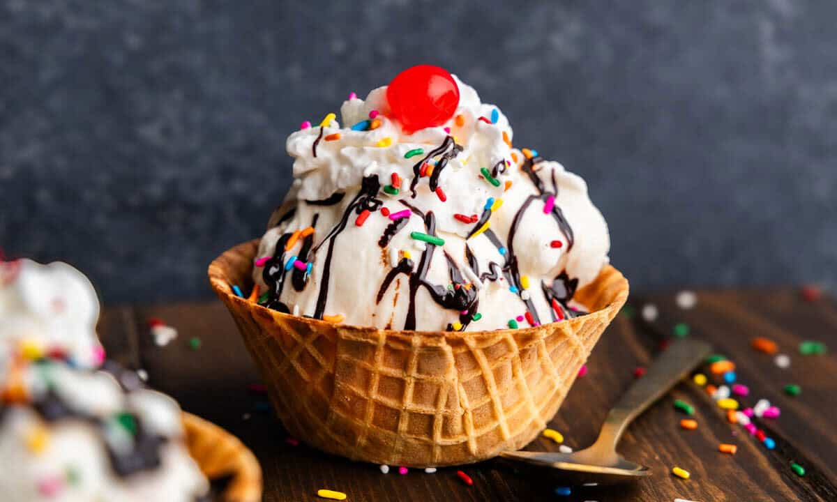 A crispy, golden-brown waffle cone overflowing with scoops of vanilla ice cream topped with multicolored sprinkles and a cherry. 